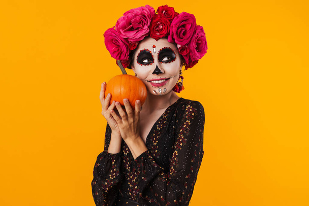Happy young girl in halloween makeup posing with pumpkin isolated over yellow background - 写真・画像