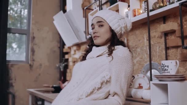 Depressed young woman, hipster, with curly hair and thick hat in the kitchen, looks unhappily at the room - Footage, Video