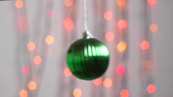 Bonito brinquedo de árvore de Natal pendura no fundo de luzes de Natal multi-coloridas e se move ligeiramente. Close-up. Brinquedo de árvore de Natal na forma de uma esfera verde. - Filmagem, Vídeo
