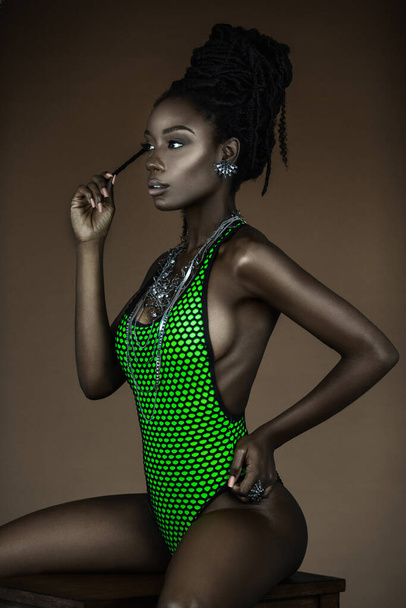 Um retrato de uma jovem mulher negra serena com longos dreadlocks, bela maquiagem e lábios úmidos posando sozinha em um estúdio com fundo marrom vestindo jóias e um maiô verde. - Foto, Imagem