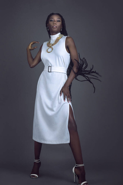 Portrait of an elegant young black female with long dreadlocks, beautiful makeup and moist lips posing by herself in a studio with grey background wearing a white dress, jewelry and high heels. - Photo, image