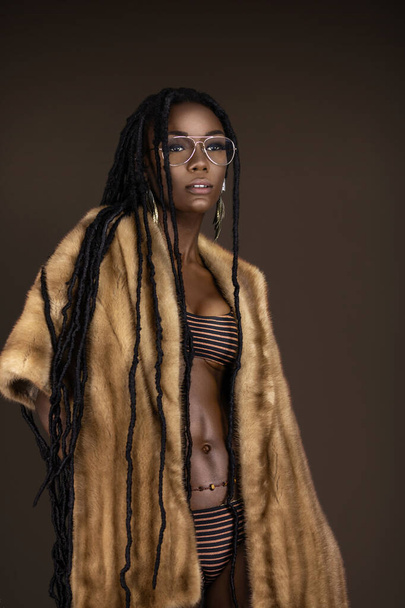 A portrait of a sexy young black female with long dreadlocks, beautiful makeup and moist lips posing by herself in a studio with dark background wearing jewelry, a bikini & fur coat. - Фото, изображение
