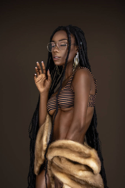 A portrait of a serene young black female with long dreadlocks, beautiful makeup and moist lips posing by herself in a studio with dark background wearing jewelry, a bikini & fur coat. - Foto, imagen