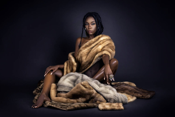 A portrait of a serene young black female with long dreadlocks, beautiful makeup and perfect lips sitting by herself on fur in a studio with dark background wearing jewelry & a fur coat. - Foto, imagen