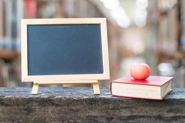 Vzdělávací koncept s prázdnou černou tabulí stojan pro oznámení s jablkem, učebnice na pozadí školní knihovny - Fotografie, Obrázek