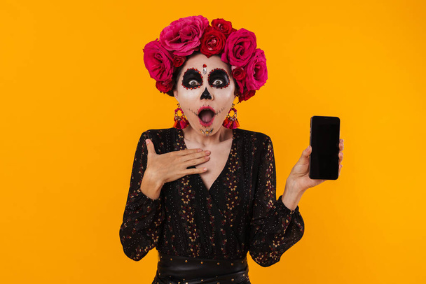 Shocked girl in halloween makeup and flower wreath showing mobile phone isolated over yellow wall - Foto, Imagen