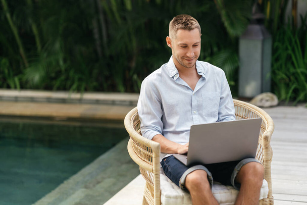 Young man using a laptop computer in a garden with a swimming pool. - Zdjęcie, obraz
