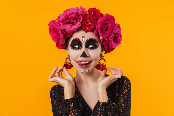 Pleased girl in halloween makeup and flower wreath showing her tongue isolated over yellow wall - Valokuva, kuva