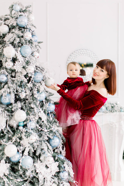 Happy young red haired mother and little daughter, wearing elegant wine red dresses, having fun and decorating christmas tree at light cozy room - Фото, зображення