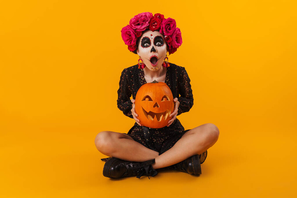Geschokt jong meisje in Halloween make-up met pompoen zitten op de vloer geïsoleerd over gele achtergrond - Foto, afbeelding