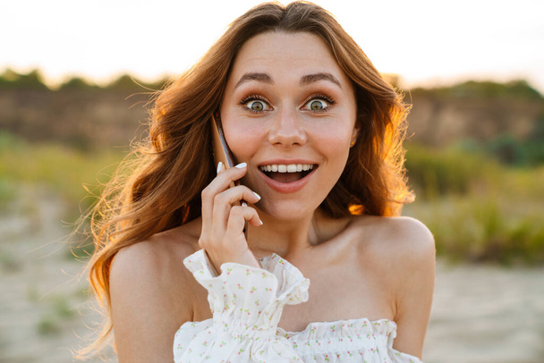 Young caucasian excited brunette woman smiling at camera while talking on cellphone outdoors - 写真・画像
