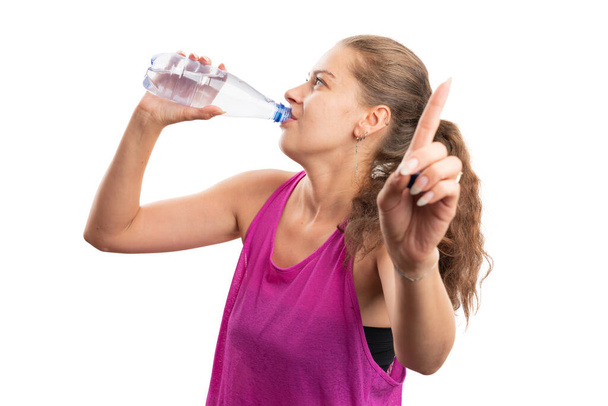Ajuste deportivo adulto hembra beber agua de la botella haciendo esperar gesto ocupado usando dedo índice usando camiseta de color rosa como traje de entrenamiento de gimnasio aislado en fondo blanco estudio - Foto, Imagen
