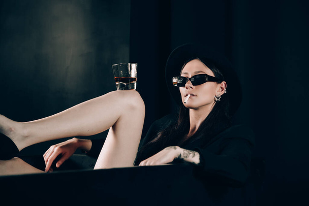 chica en una chaqueta negra y sombrero fumar cigarrillo posando cerca de un baño negro. Un vaso con alcohol en la mano. Elegantes gafas de sol. Largas piernas sexy desnuda - Foto, Imagen