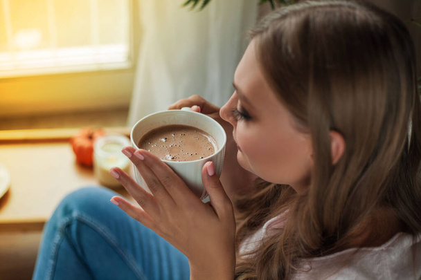 Beautiful young woman drinking hot cocoa and sitting by the window at home - Φωτογραφία, εικόνα
