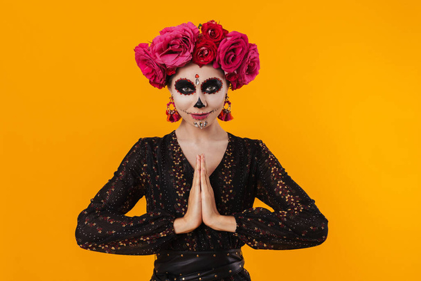 Pleased girl in halloween makeup and flower wreath meditating on camera isolated over yellow wall - Fotoğraf, Görsel