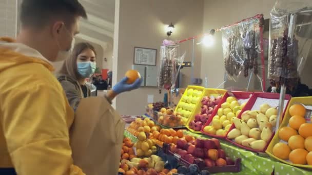Koruyucu maskeli ve eldivenli genç bir çift taze portakalları seçiyor ve onları bir kese kağıdına koyup pazarda satıcısına veriyor. - Video, Çekim