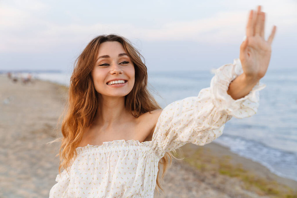 Young caucasian attractive brunette woman in dress smiling and waving on beach - 写真・画像