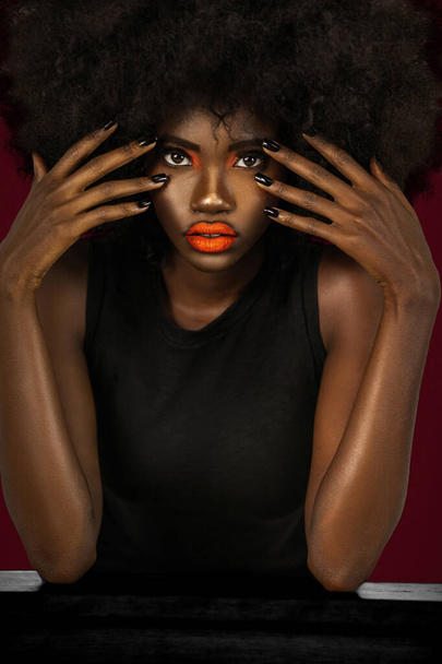 Portrait of a flirtatious young black female with long black hair, popping orange lip stick, perfectly manicured nails is wearing a big afro & leaning on a table in front of a dark red background. - Fotografie, Obrázek