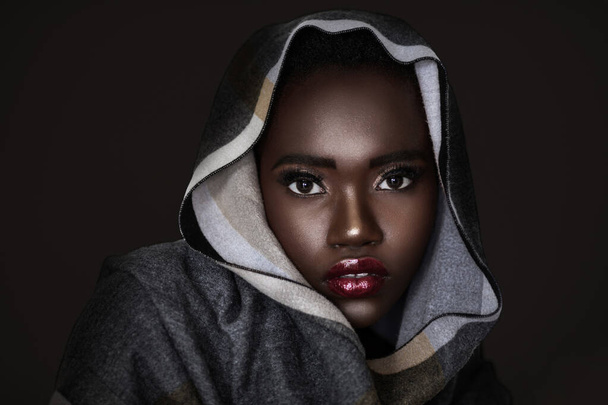 Ein Kopfschuss einer netten jungen schwarzen Frau mit kurzen schwarzen lockigen Haaren, schönem Make-up und leuchtend rotem Lippenstift, die ein buntes Kopftuch vor schwarzem Hintergrund trägt. - Foto, Bild