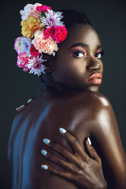 Un retrato de una sexy joven hembra negra con un afro corto, hermoso maquillaje y labios húmedos posando sola en un estudio con fondo oscuro con un ramo de flores en el pelo. - Foto, imagen