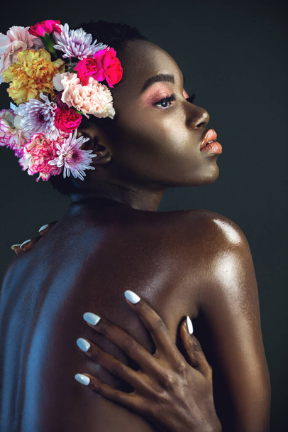 短いアフロ、美しいメイクと湿った唇を持つ穏やかな若い黒人女性の肖像画彼女の髪に花の花束を身に着けている暗い背景を持つスタジオで自分自身によってポーズ. - 写真・画像
