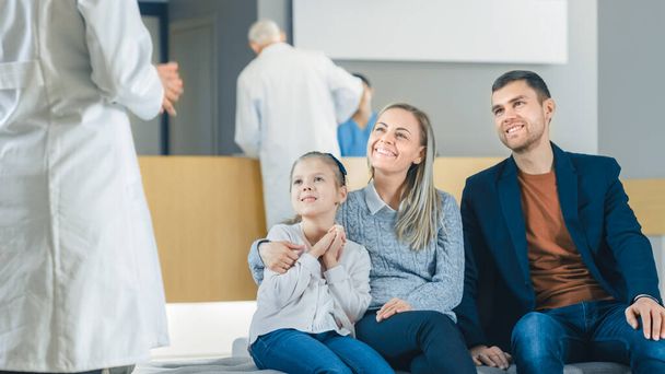 In the Hospital, Doctor Approaches Young Family of Father, Mother and Cute Little Daughter Sitting in the Lobby. The News are Good, Happy Moment in the Life of Young Family. Modern, Clean Hospital. - 写真・画像