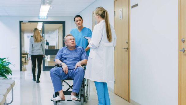 In the Hospital Hallway, Nurse Pushes Elderly Patient in the Wheelchair, Doctor Talks to Them while Using Tablet Computer. Clean, New Hospital with Professional Medical Personnel. - Photo, Image