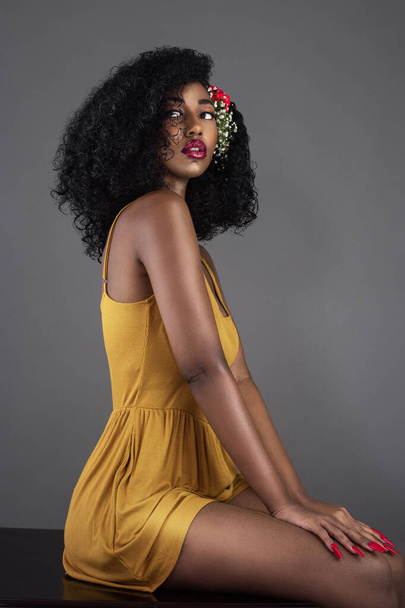 A portrait of a serene young black female with curly long black hair, beautiful makeup and red lips sitting on a desk by herself in a studio wearing a yellow dress & flowers in her hair. - Photo, Image