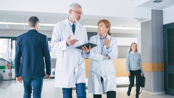 In the Hospital, Busy Doctors Talk, Using Tablet Computer While Walking Through the Building. In the Background Patient Talks with Receptionist. New Modern Fully Functional Medical Facility. - 写真・画像