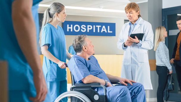 In the Hospital Lobby, Nurse Pushes Elderly Patient in the Wheelchair, Doctor Talks to Them while Using Tablet Computer. Clean, New Hospital with Professional Medical Personnel. - Foto, Bild