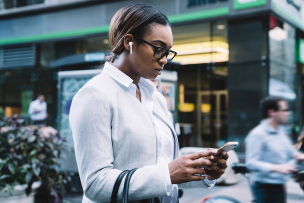 Concentrated African American female in white jacket and stylish glasses listening to music and browsing smartphone while walking on city street - Fotó, kép