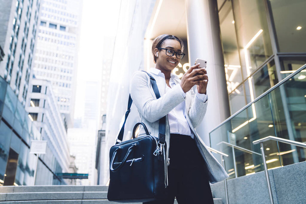 From below of happy content black woman with wireless earphones  surfing smartphone in downtown while walking among office buildings - Foto, Imagen
