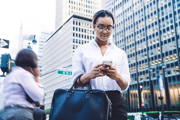 Mulher executiva afro-americana inteligente satisfeita jovem em óculos e com saco segurando celular enquanto está em pé nas ruas da cidade movimentadas modernas - Foto, Imagem