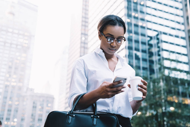 Black busy serious brunette in glasses messaging on mobile phone carrying black bag and holding cup of coffee with modern city on background - Foto, Imagem