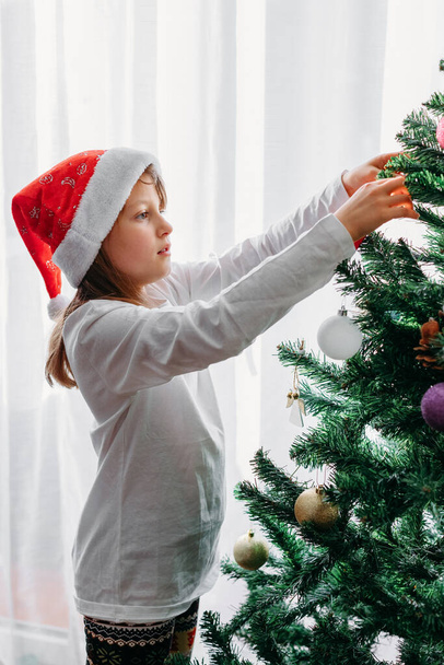 Мила дівчина з темним волоссям у капелюсі Санта прикрашає дерево новими роками, різдвяними іграшками. Присвячення для нового року і Різдва Христового. Вертикальна орієнтація - Фото, зображення