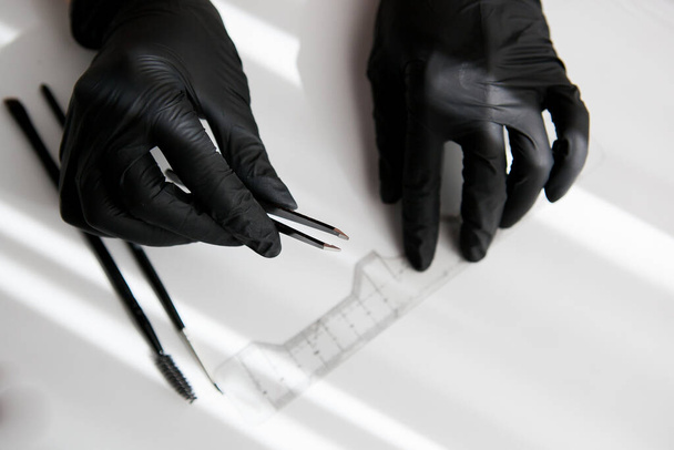 the master in black vinyl gloves holds a pair of tweezers for making eyebrows on the background of a ruler, brushes. - Foto, Imagem