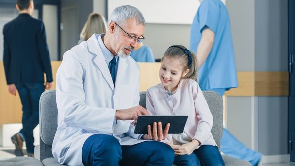 In the Lobby of the Hospital Male Doctor Talks with a Cute Little Girl while Sitting on the Sofa, Hes Showing Tablet Computer to Her. Busy Modern Hospital with Best Possible Pediatrics Department in - Foto, Bild