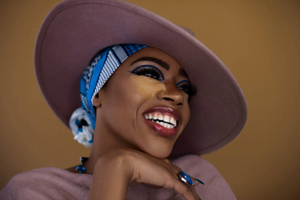 Uma bela jovem mulher negra com um lindo sorriso e maquiagem posando sozinha dentro de um estúdio usando um chapéu cinza, lenço de cabeça azul e jóias. - Foto, Imagem