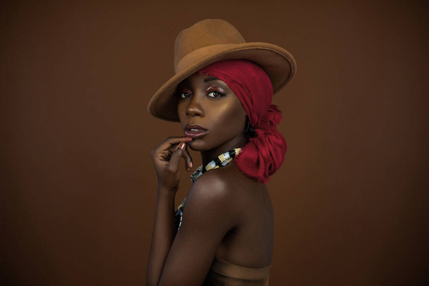 Zmysłowo wyglądająca młoda czarna kobieta z pięknym makijażem, pozująca sama w studio w brązowej bluzce bez ramiączek, czerwonej chustce na głowie, brązowym kapeluszu i wspaniałym kolorowym naszyjniku. - Zdjęcie, obraz
