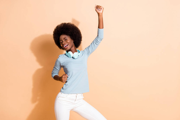 Foto retrato de niña afroamericana despreocupada bailando con un puño en el aire aislado sobre fondo de color beige pastel - Foto, Imagen