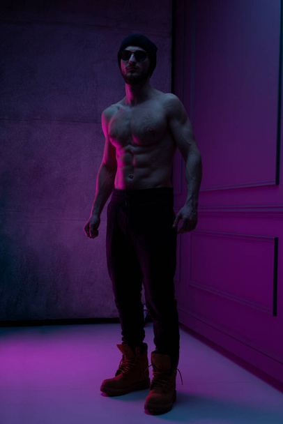 Ganzkörperporträt eines brutalen Mannes mit nacktem Oberkörper, der im Studio mit Betonwand in lila Neonfarbe posiert. - Foto, Bild