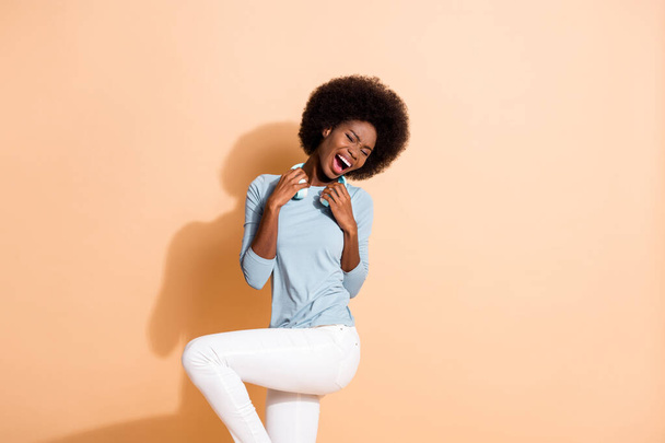 Photo portrait of wild african american woman holding headphones with two hands dancing standing on one leg isolated on pastel beige colored background - Foto, afbeelding