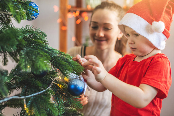 Mother with her child son is decorating the Christmas tree with toys and Christmas balls. Happy family time on new year's eve at home - Фото, зображення