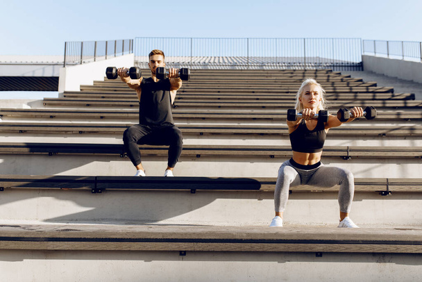 Sporty couple in sportswear doing squats with dumbbells outdoors on city stairs - Foto, imagen