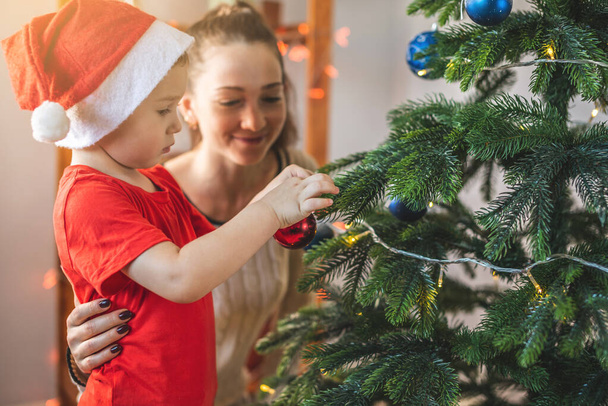 Mother with her child son is decorating the Christmas tree with toys and Christmas balls. Happy family time on new year's eve at home - Foto, Bild