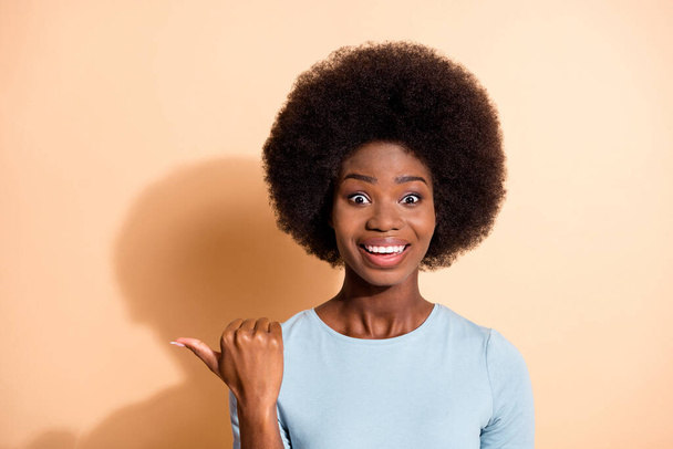 Photo portrait of excited african american girl pointing thumb at blank space isolated on pastel beige colored background - Фото, изображение