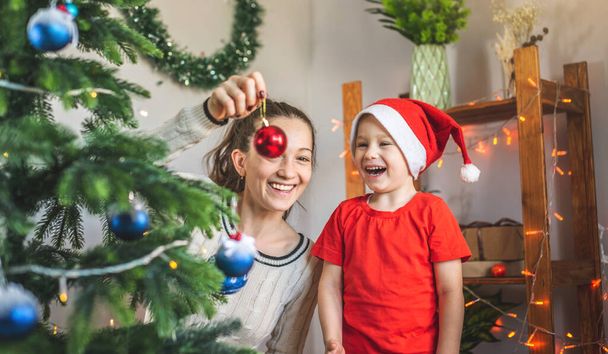 Mother with her child son is decorating the Christmas tree with toys and Christmas balls. Concept of a happy loving family on new year's eve - Фото, изображение