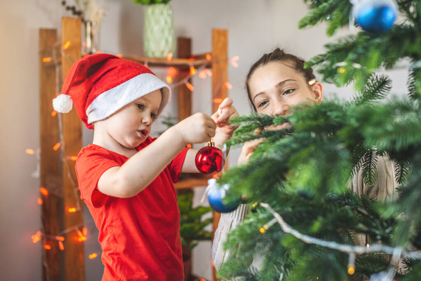 Mother with her child son is decorating the Christmas tree with toys and Christmas balls. Concept of a happy loving family on new year's eve - Zdjęcie, obraz