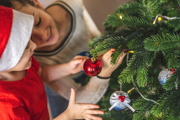 Mother and her child son are holding a Christmas tree ball toy to hang on a branch. The concept of a happy loving family on new year's eve - Фото, изображение