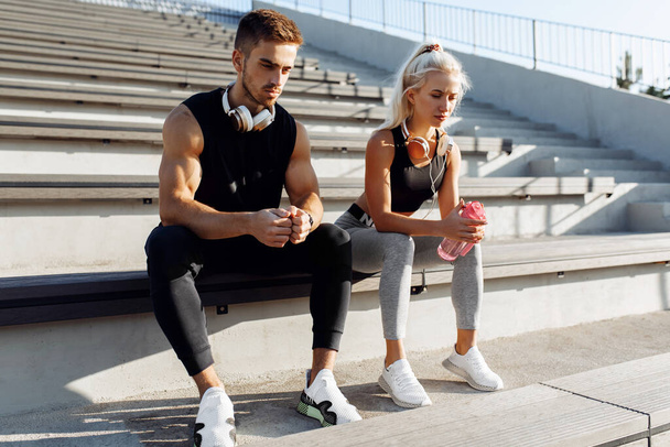 Young sportive couple in sportswear, taking a break to drink water, sitting on the stairs outdoors, healthy lifestyle, sport - Photo, Image